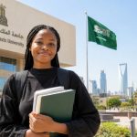 Student holding books in front of a university building with the Saudi flag